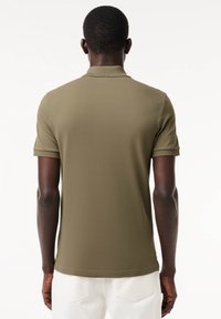 Homme à la peau foncée portant un polo vert olive à manches courtes et un pantalon blanc, vu de dos sur un fond blanc.