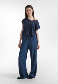 Młoda kobieta stojąca z odsłoniętym jednym ramieniem, ubrana w luźną granatową bluzkę, szerokie dżinsowe spodnie i czarne buty na białym tle.