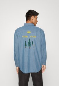 Light blue denim shirt featuring an embroidered design on the back with yellow sun, green trees, and "GONE HIKING" text. Long-sleeve with button cuffs.