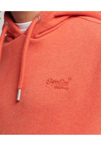 Sudadera de color coral con cordones, que presenta un logo bordado de "Superdry" en color a juego; confeccionada con un tejido suave y texturizado. Aglets plateados en los cordones.