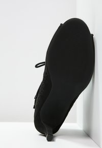 Zapato de tacón alto de ante negro que presenta una puntera afilada y un tacón delgado; cuenta con una plantilla texturizada y una suela de goma lisa.