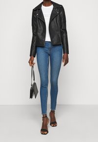 Giacca da biker in pelle nera con dettagli in metallo argento, camicia bianca texturizzata, jeans skinny blu e sandali neri con cinturini, che tiene in mano una borsa nera piccola.