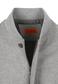 Grijze wollen jas met zilveren rits gedeeltelijk open en zwarte knop bij de kraag, met een oranje label aan de binnenkant.