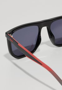 Ochelari de soare negri cu lentile închise, având un design îndrăzneț și unghiular și brațe cu dungi roșii. Accentele includ un logo pe lateral.