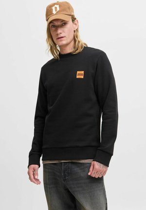Black sweatshirt with ribbed cuffs and hem. Features an orange square logo. Paired with dark jeans and a brown cap with white detailing.