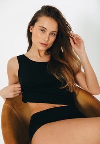 Sort, tætsiddende tanktop med bred halsudskæring og blød tekstur, parret med matchende højtaljede shorts, siddende i en tanfarvet stol.