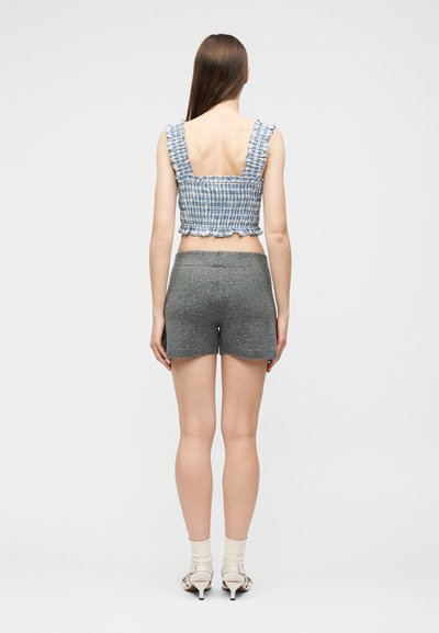 Γυναίκα με μακριά καστανά μαλλιά, να φορά ένα μπλε καρό αμάνικο κοντό τοπ, γκρι σορτς με σπίθες, λευκές κάλτσες και λευκές γόβες με μικρό τακούνι, όρθια από πίσω.