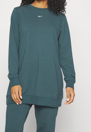 Personne portant un sweat-shirt Nike bleu sarcelle à manches longues et un pantalon assorti, debout, les mains détendues le long du corps sur un fond uni.