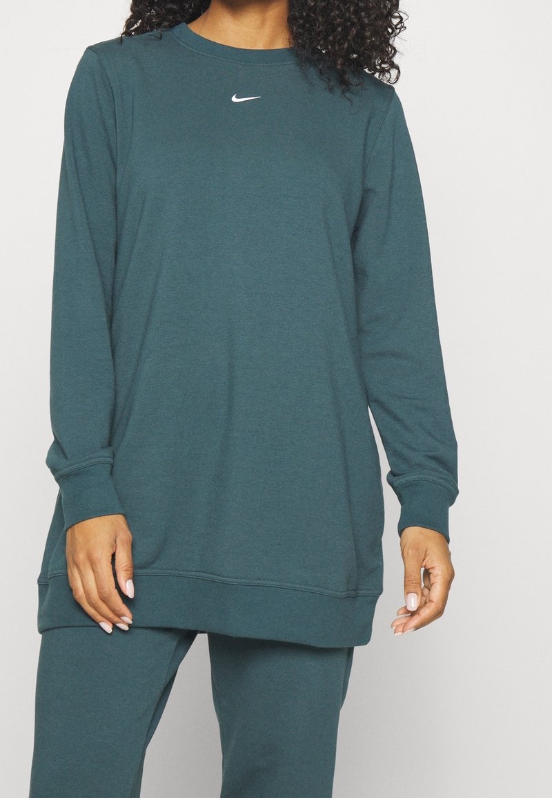 Personne portant un sweat-shirt Nike bleu sarcelle à manches longues et un pantalon assorti, debout, les mains détendues le long du corps sur un fond uni.