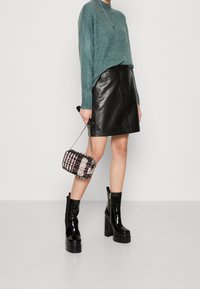 Un pull tricoté teal, une jupe en cuir noire, des bottes à plateforme noires brillantes. Tenant un mini sac à main rose et noir à carreaux avec une chaîne.