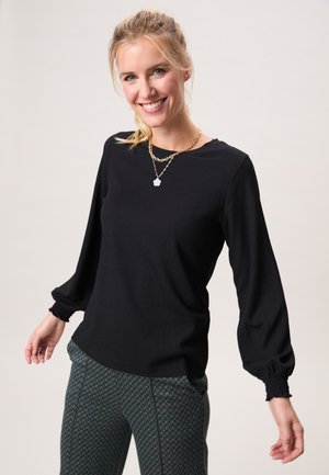 Lachende vrouw met blond haar in een paardenstaart, gekleed in een zwarte blouse met lange mouwen, patroonbroek en een gouden ketting met een hanger.