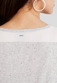 Närbild av en persons nacke och axel som bär en prickig ljusgrå topp med en liten "RAW"-logotyp under halsringningen och ett stort silverfärgat hoopsmycke.