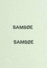 Tissu en maille vert clair avec deux logos noirs "SAMSØE" imprimés verticalement. Texture lisse et design minimaliste.
