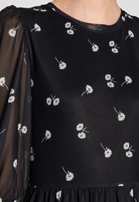Schwarzes Blumentop mit einem schwarzen, transparenten Puffärmel, kleinen weißen Gänseblümchenmustern und einem gerafften Saum. Glattes Material mit leichter Textur.