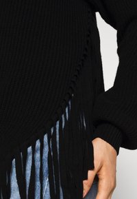 Närbild av hand i jeansficka, iklädd en svart ribbad tröja med fransdetaljer på sidan och ballongärmsmudd.