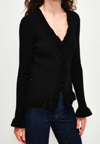 Czarny prążkowany kardigan z długim rękawem, z falbankami na dole i przy dekolcie. Charakteryzuje się dopasowanym krojem i delikatnie rozszerzanymi mankietami. Połączony z ciemnym dżinsem.