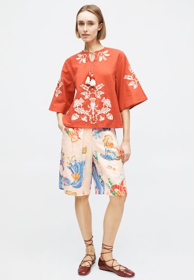 Femme portant un chemisier orange brodé aux larges manches, un short mi-cuisses à motif multicolore, et des chaussures plates marron à lacets.