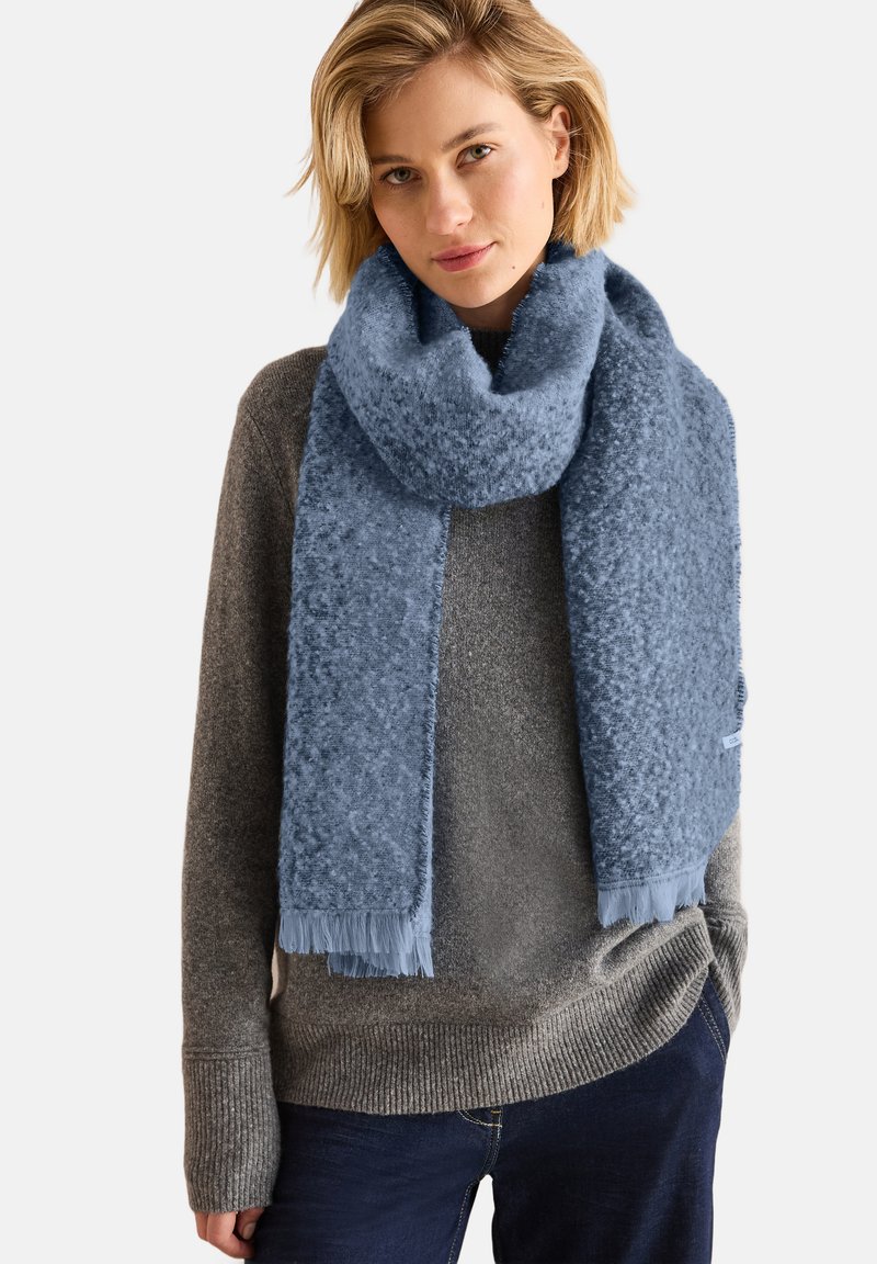 Bufanda azul clara hecha de un suave tejido texturizado con bordes desflecados. Llevada suelta alrededor del cuello, contra un fondo de suéter gris.