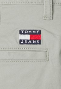 Tessuto di cotone grigio chiaro con una piccola tasca, caratterizzato da un'etichetta ricamata rettangolare con "TOMMY" e "JEANS" in bianco e blu, con accenti rossi.