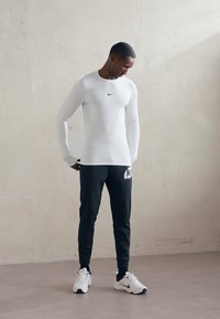 Mies, który ma na sobie biały długi rękaw Nike koszulka kompresyjna, czarne dresy Nike i białe buty Nike, stoi pod jasną ścianą.