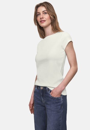 Junge Frau mit hellbraunen Haaren, die ein kurzärmliges, cremefarbenes Oberteil und blaue Jeans trägt, steht mit einer Hand in der Tasche.