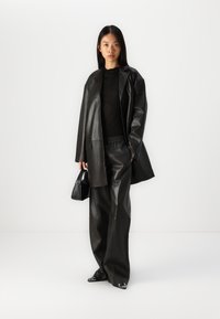 Zwarte leren oversized blazer gecombineerd met losse leren broeken. Model houdt een kleine zwarte handtas vast; hoge, puntige zwarte schoenen.