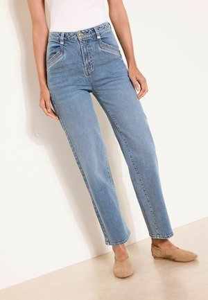 Henkilö, joka pukeutuu vaaleansinisiin korkeavyötäröisiin farkkuihin ja beigeihin liukukengiin, seisoo valkoisen seinän ja vaalean lattian edessä.