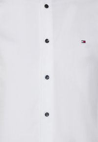 Camicia bianca con bottoni neri, dotata di sei bottoni neri e un piccolo logo ricamato in blu, rosso e bianco sul lato sinistro del petto. Tessuto liscio.