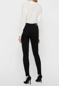 Jeans negros ajustados con una textura suave, cintura alta y passadores para cinturón. Combinados con una camisa blanca de botones. Zapatos negros de tacón alto.