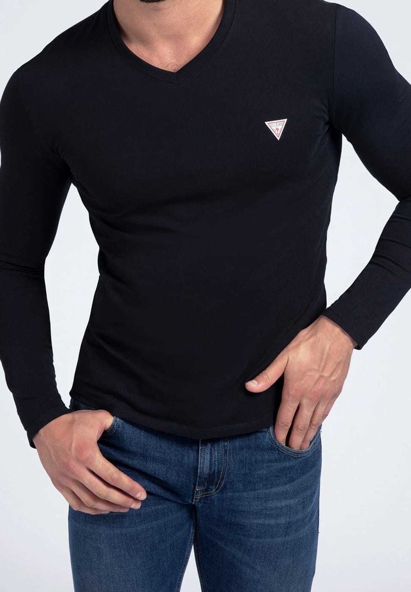 Uomo che indossa una maglietta nera attillata a maniche lunghe con scollatura a V e un piccolo logo triangolare bianco sul petto, abbinata a jeans in denim blu.