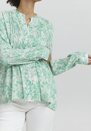 Kvinde iført en hvid bluse med grønne blomsterprint og plisseringer, kombineret med hvide bukser, foran en ensfarvet lys baggrund.