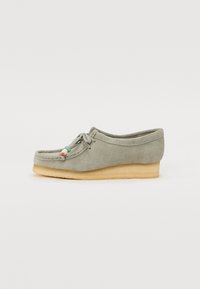 Mocassin en velours gris avec un petit talon, un nez arrondi et un laçage décoratif agrémenté de perles colorées. Texture douce avec un design minimaliste.