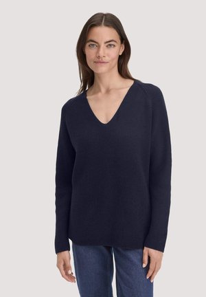 Chunky marineblauwe gebreide trui met een diepe V-hals, lange mouwen en ribbelstructuur, gecombineerd met denimjeans. Eenvoudig, ontspannen ontwerp.