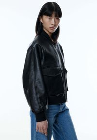 Schwarze Lederbomberjacke mit übergroßen Ärmeln, zwei Fronttaschen, gerippten Bündchen und einem Reißverschluss, kombiniert mit blauen Jeans.