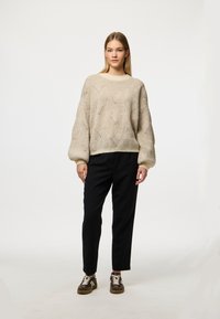 Lys beige oversized genser med diamantstrikkemønster, ribbestrikkede mansjetter og hals, sammen med svarte bukser og brune sneakers.