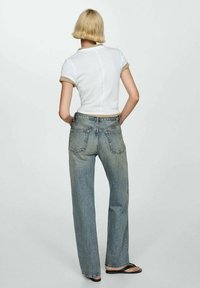 T-shirt bianca aderente in cotone con bordi beige, abbinata a jeans a vita alta di colore blu chiaro. I jeans presentano tasche posteriori classiche e gamba dritta.