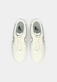 Białe sneakersy Nike Air z czarnymi akcentami, okrągły czubek, perforowana górna część dla wentylacji, płaskie sznurowadła i szara podszewka.