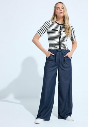 Donna che indossa una maglietta a maniche corte a righe bianca e nera, pantaloni navy a gamba larga con tasche e sneakers bianche, in piedi con le mani nelle tasche.