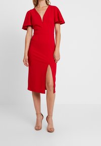 Czerwona dopasowana sukienka z głębokim dekoltem w kształcie V i falbaniastymi rękawami. Posiada wysokie rozcięcie na lewym boku, wykonana z gładkiego materiału.