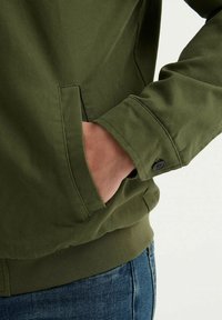 Veste en coton vert avec une finition texturée, dotée d'une poche sur le côté gauche et de détails de poignets boutonnés. Portée avec un jean en denim bleu.