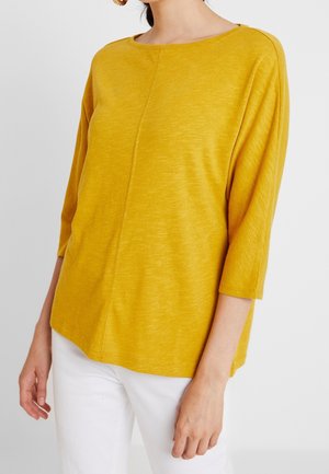 Femme portant un haut jaune moutarde à manches trois-quarts associé à un pantalon blanc, sur un fond clair uni.