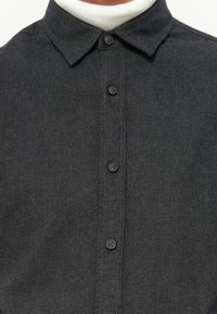 Camicia button-up grigio scuro con colletto classico, dotata di bottoni neri e tessuto testurizzato. In primo piano, evidenzia la pattina e le cuciture.