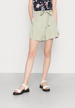 Persona lleva shorts de talle alto verde salvia con cinturón atado, top negro de flores y sandalias blancas, de pie sobre un fondo neutro.