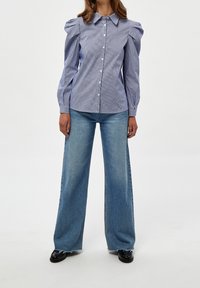 Blusa a rayas con mangas abullonadas, cierre frontal de botones y cuello. Combinada con jeans de mezclilla azul de pierna ancha y zapatos negros.
