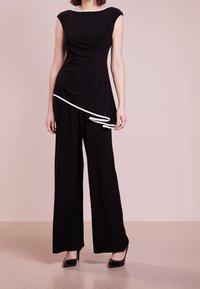 Svart ärmlös jumpsuit med draperad design, med vit kant längs halsringningen och midjan, samt vida byxor. Slät tygtextur.