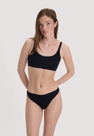 Frau mit langen braunen Haaren, die einen schwarzen Sport-BH und schwarze Bikini-Unterwäsche trägt, steht vor einem schlichten hellen Hintergrund.