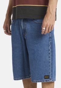 Mann trägt mittelblaue Jeansshorts und ein schwarz-braunes Hemd, mit silbernen Ringen an beiden Händen.