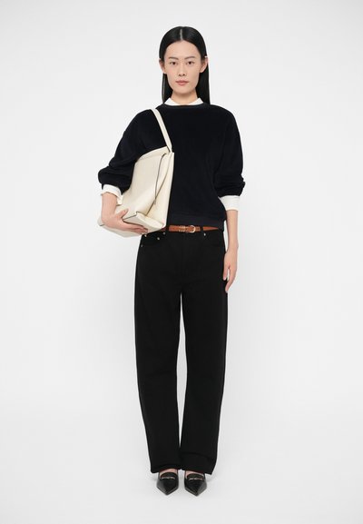 Hosszú, egyenes fekete hajú nő fekete pulóverben, fehér ingujjakkal, fekete bő szárú nadrágban, fekete hegyes orrú cipőben, bézs színű tote táskát tartva.