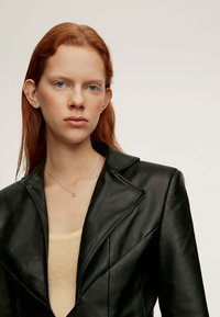 Blazer de cuero negro con solapa con muesca y picos afilados. Usado sobre un suéter de color beige claro. La modelo tiene el pelo rojo y joyas sutiles.