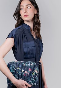 Borsa trapuntata blu navy con un motivo floreale che presenta vari colori. Tessuto liscio con una forma arrotondata, tenuta da una persona in un vestito scuro.